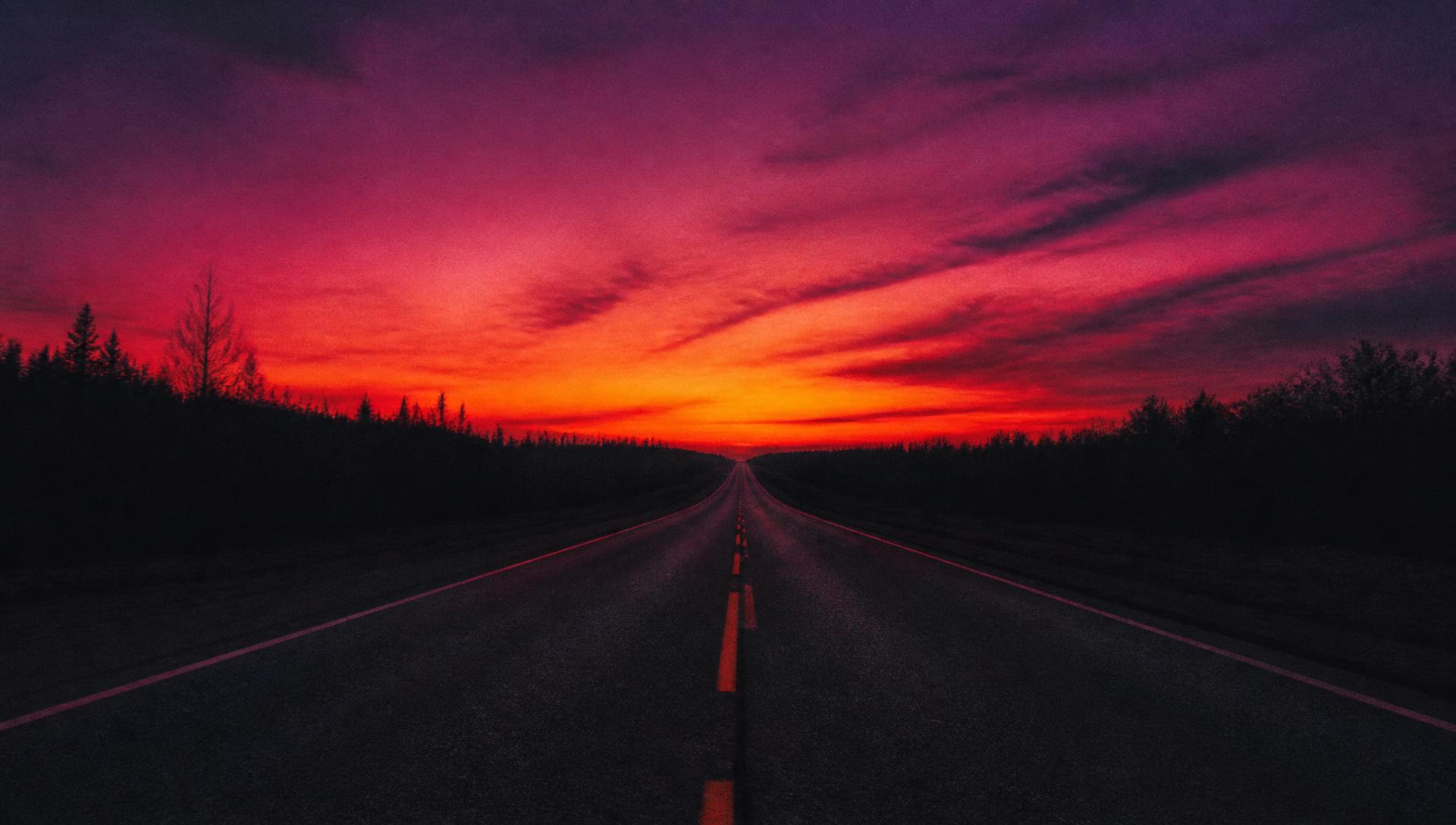 Open Michigan highway stretching toward an orange and magenta sunset, evoking Travis Hermiz's road-trip inspired brand aesthetic